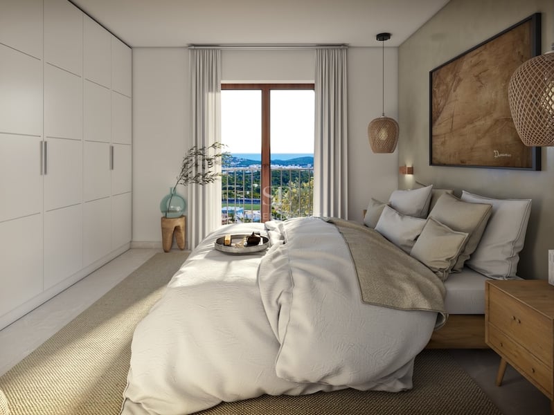 Bright master bedroom with a large bed, built-in white wardrobes, and a window overlooking a coastal landscape.