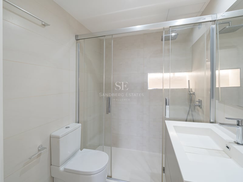 Ljust modernt badrum med en dusch i glas, vit toalett och minimalistiskt tvättställ.