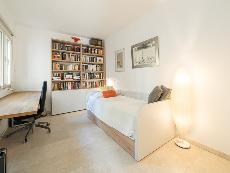 Ljust sovrum med enkelsäng, stor bokhylla i trä, skrivbord och en stämningsfull golvlampa.