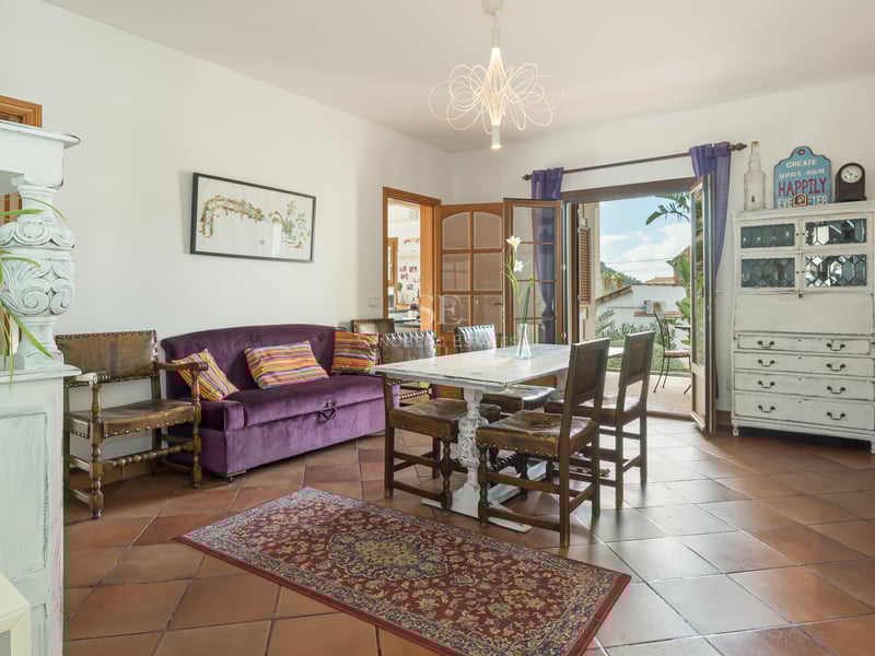 Dining room with terracotta floors, purple velvet sofa, and white wooden table opening to a sunny balcony.