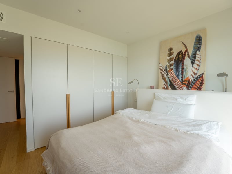 Ljust sovrum med vit säng, stora inbyggda garderober med trädetaljer och en tavla med fjädrar.