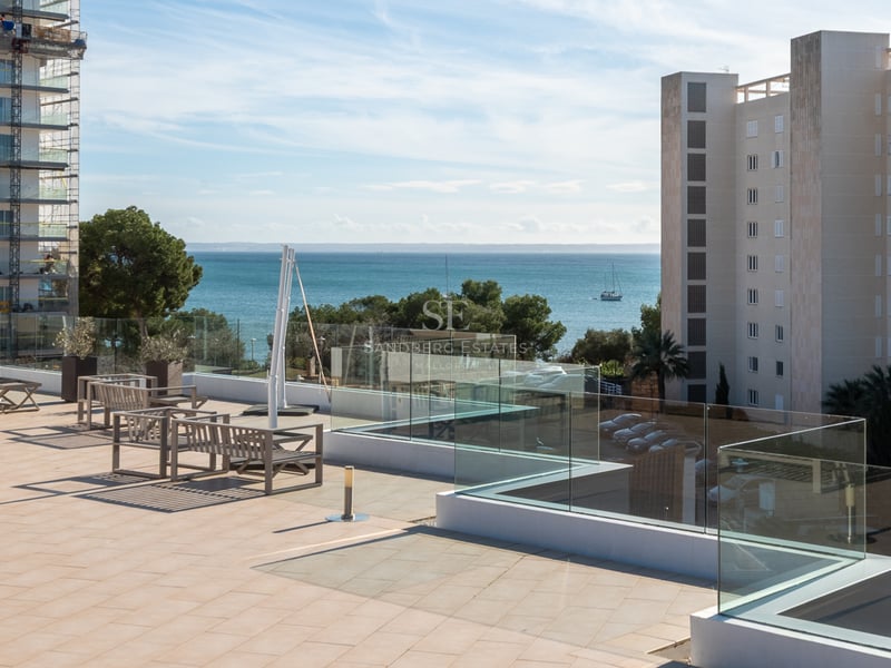 Grande terrasse carrelée avec garde-corps en verre et meubles en bois, surplombant la mer bleue et les bâtiments.