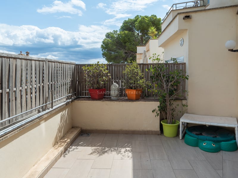 Terrasse privée carrelée avec clôture en bois, plantes en pots et vue sur la végétation sous un ciel bleu.