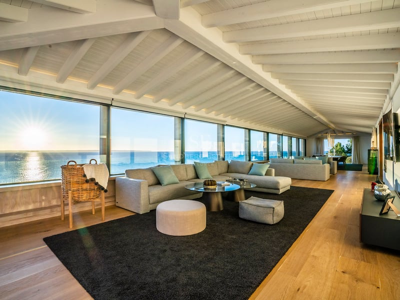 Modernes Wohnzimmer mit weißen Deckenbalken, Eichenparkett und bodentiefen Fenstern mit Meerblick bei Sonnenuntergang.