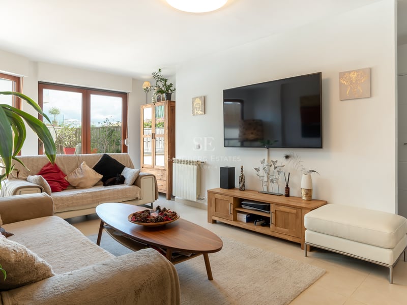 Salon lumineux avec canapés beiges, meubles en bois, grand téléviseur mural et accès à une terrasse extérieure.