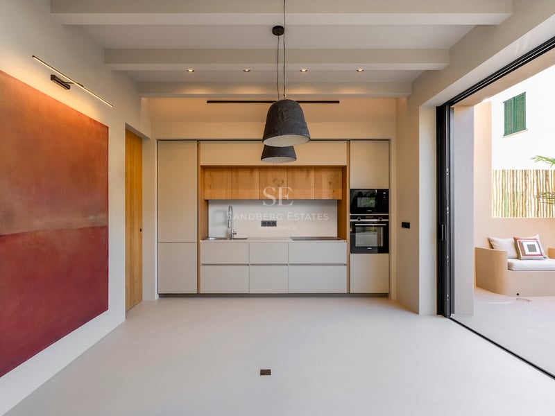 Cuisine minimaliste avec accents en bois clair, grand tableau rouge et baies vitrées ouvrant sur un patio ensoleillé.