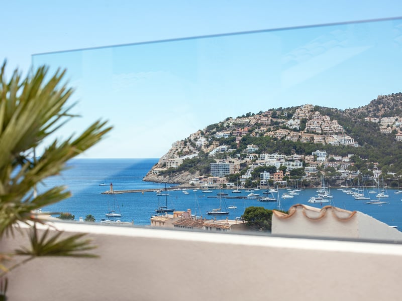 Vue sur un port méditerranéen avec bateaux et villas à flanc de colline à travers un garde-corps en verre.