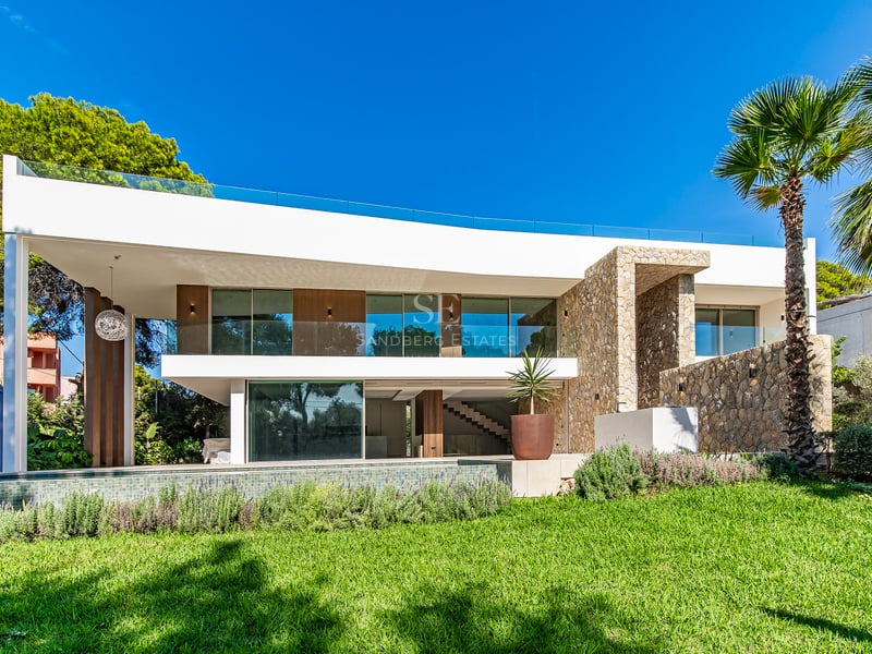 Villa moderne blanche de deux étages avec murs en pierre, balcons en verre et piscine donnant sur une pelouse verte.