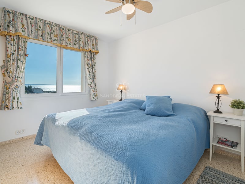 A bright bedroom with a blue bedspread, sea views through a floral-curtained window, and bedside lamps.