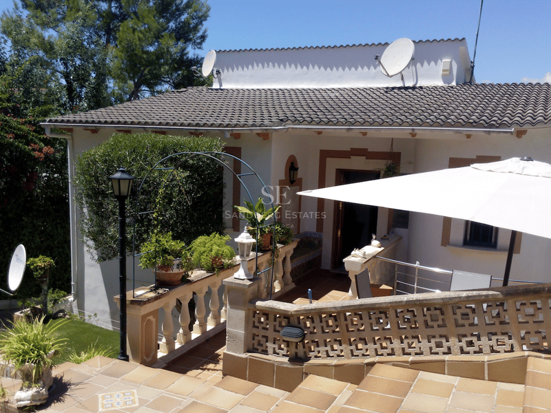 Vitkalkad villa med trappor i terrakotta, dekorativa stenräcken och ett parasoll under en klarblå himmel.