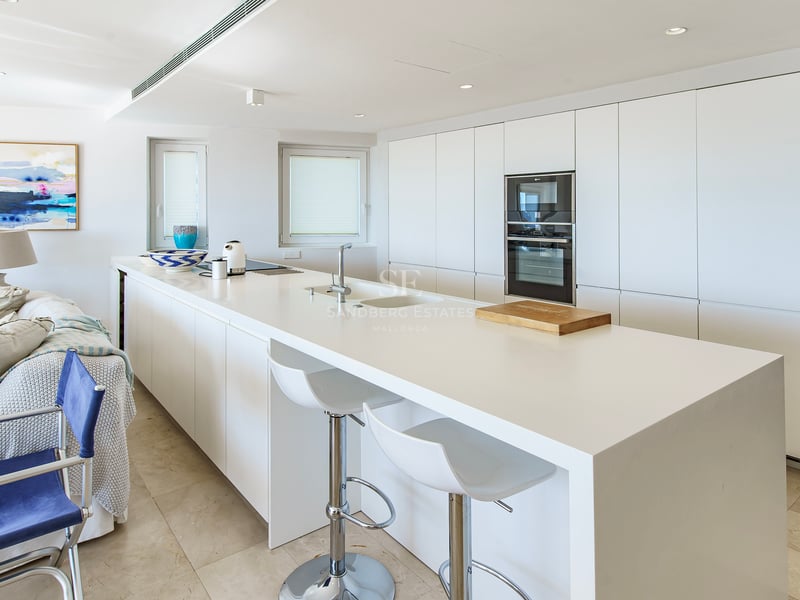 A bright, modern white kitchen featuring a large central island with bar stools and integrated appliances.