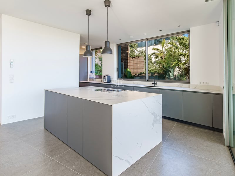 Cuisine moderne avec un grand îlot en marbre blanc, des meubles gris et une fenêtre donnant sur un jardin tropical.