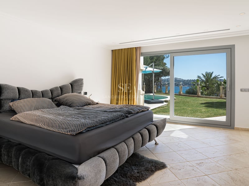 Modernes Schlafzimmer mit grauem Polsterbett und großen Glasschiebetüren, die zum Garten, Pool und Meer führen.