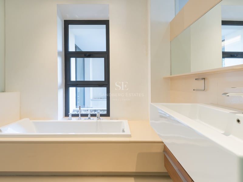 Modernt badrum med ett inbyggt vitt badkar intill ett fönster och ett stort rektangulärt tvättställ med stor spegel.