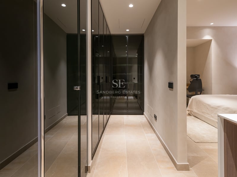 Un couloir avec des armoires en verre teinté du sol au plafond menant à une chambre moderne avec éclairage encastré.