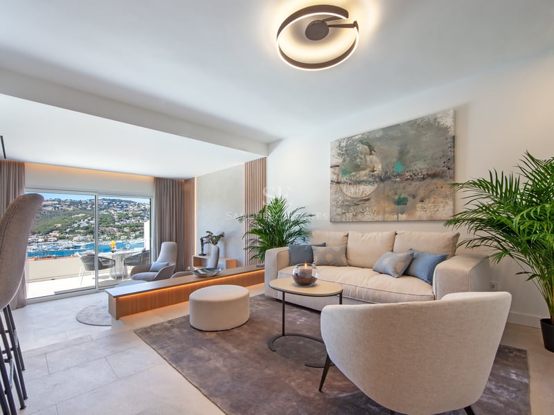 Salon moderne et lumineux avec mobilier crème, grandes baies vitrées sur la mer et plantes vertes.