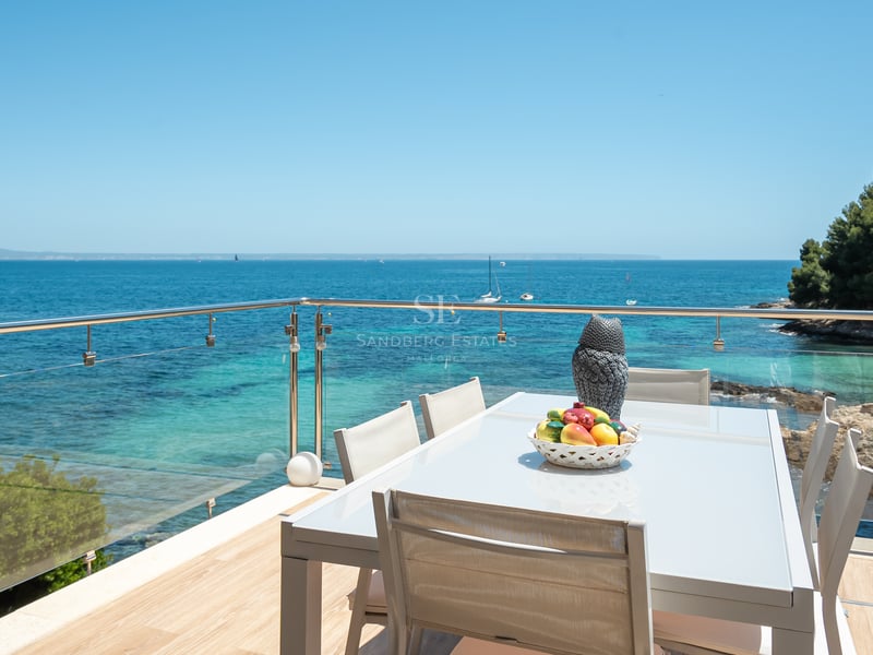 Modern matplats utomhus på en terrass med glasräcke och utsikt över en turkos vik och klarblå himmel.