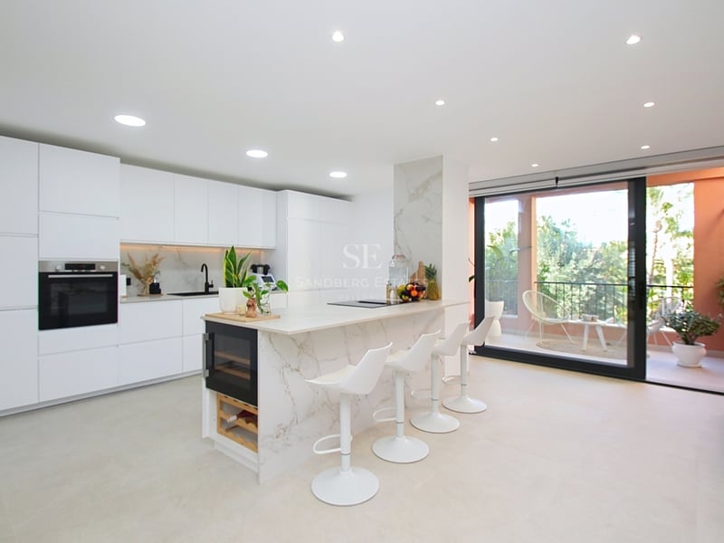 Minimalistiskt vitt kök med marmorö, vinkyl och glaspartier mot terrass.