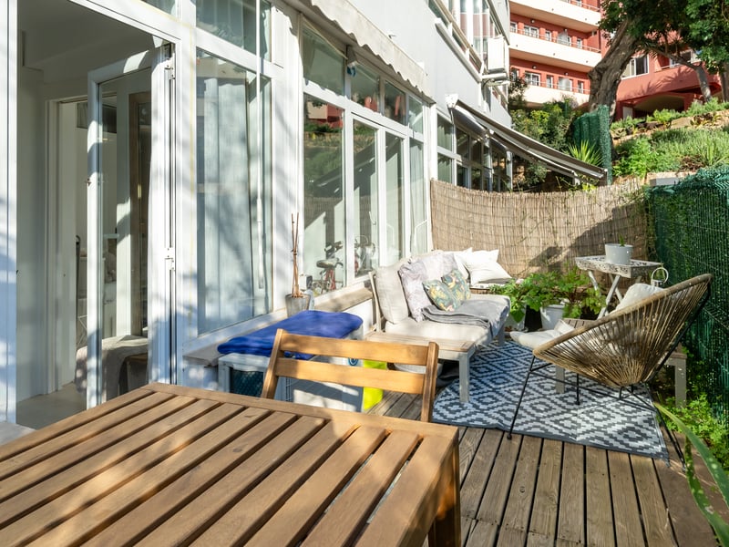 Private wooden terrace with outdoor lounge furniture, geometric rug, and glass sliding doors under bright daylight.