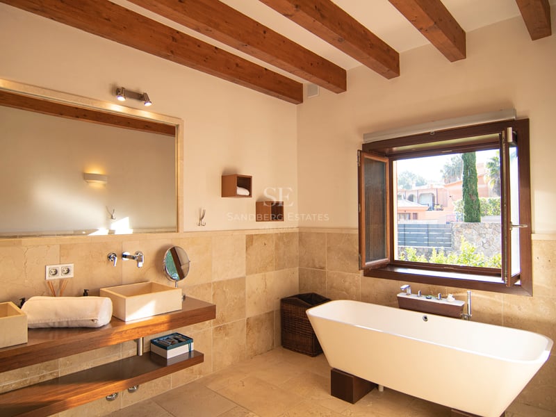 Badrum med synliga takbjälkar, fristående vitt badkar, dubbla stentvättställ och fönsterutsikt.