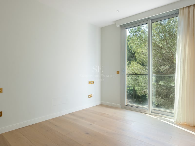 Empty modern bedroom featuring light oak floors, white walls, and large sliding glass doors overlooking green trees.