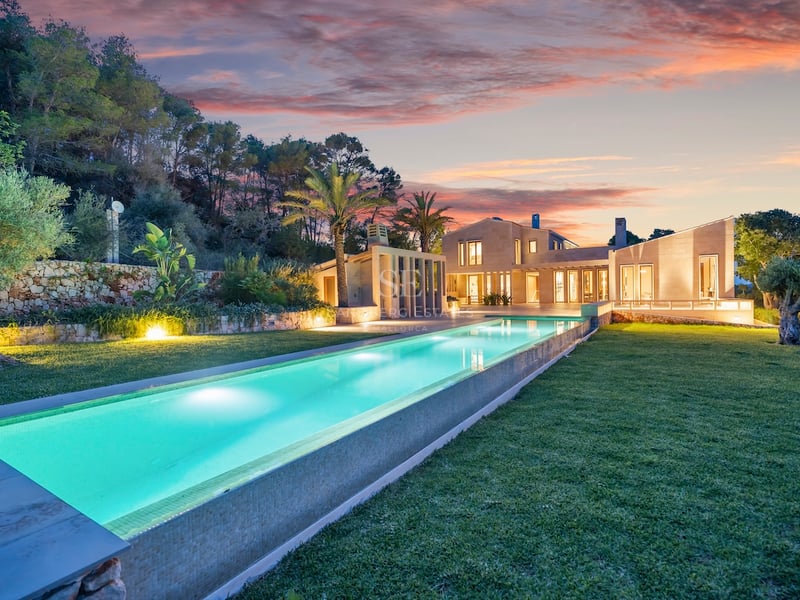 Longue piscine illuminée menant à une villa moderne en pierre sous un ciel de coucher de soleil éclatant.
