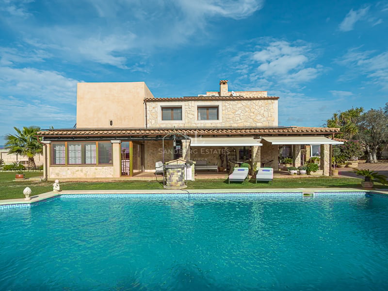 Grande piscine turquoise devant une villa méditerranéenne en pierre avec terrasse couverte et jardins.