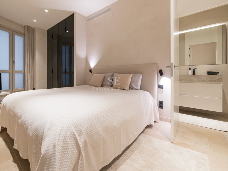 Chambre principale moderne avec textiles crème, sol en pierre, placards sombres et salle de bain attenante.