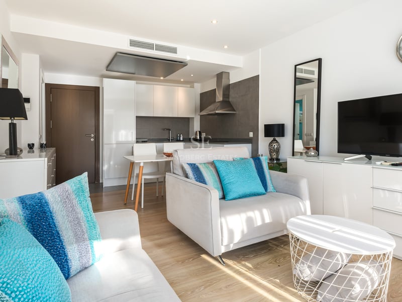 Salon lumineux avec fauteuil gris, coussins turquoise, meuble TV et cuisine moderne en arrière-plan.