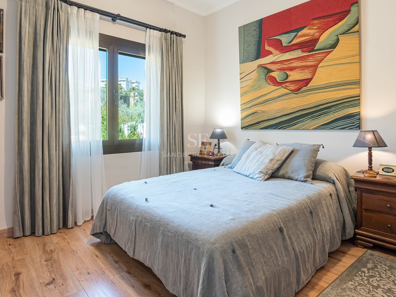 Ljust sovrum med trägolv, säng i gråa toner, klassiska nattduksbord och en stor färgstark tavla.