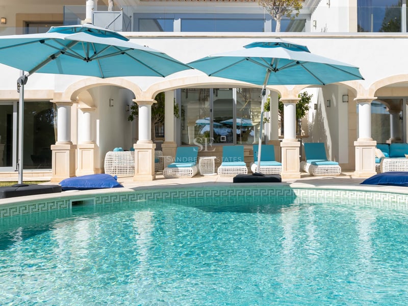 Türkisfarbener Pool mit klassischen weißen Säulen und blaugrünen Sonnenschirmen auf einer sonnigen Terrasse.