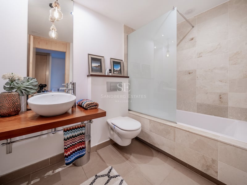 Salle de bain moderne avec plan de toilette en bois, vasque à poser, WC suspendu et baignoire carrelée en pierre.