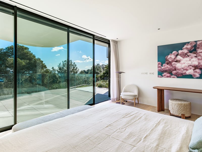 Chambre lumineuse avec lit blanc, baies vitrées du sol au plafond et grande terrasse avec vue sur les arbres.