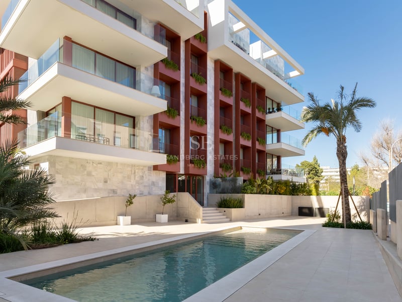 Modern apartment building facade with glass balconies and a rectangular swimming pool framed by palm trees.