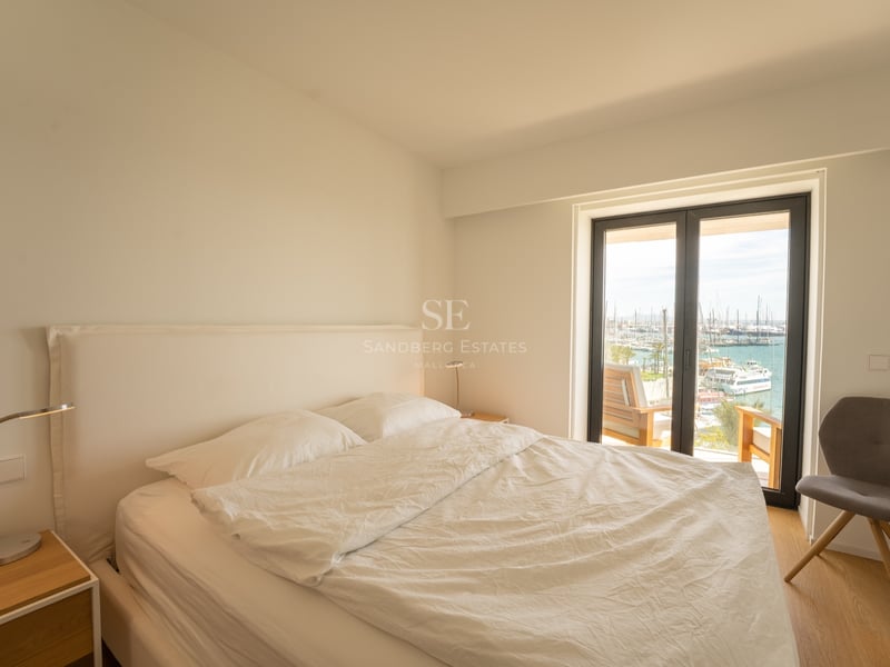 Ljust sovrum med vit säng, grå stol och stora glasdörrar mot en marina fylld med båtar.