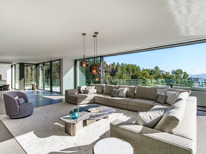 Grand salon moderne avec baies vitrées du sol au plafond, canapé gris en L et vue sur la forêt.