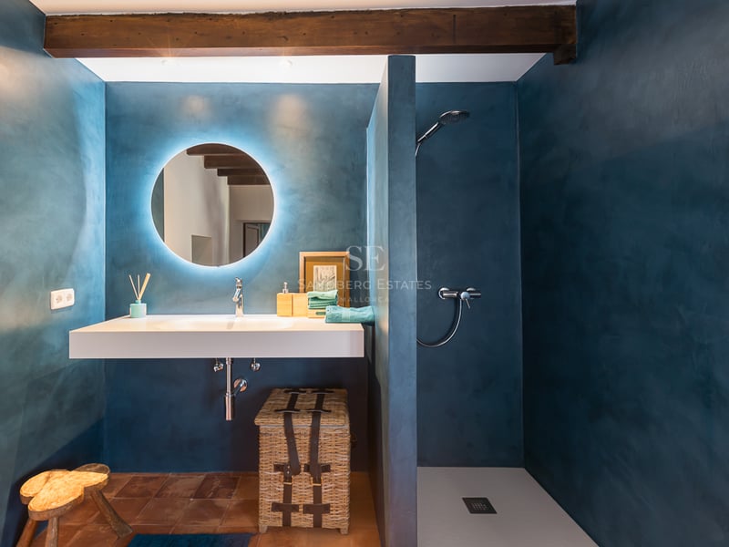 Salle de bain moderne avec murs en microciment bleu, miroir circulaire rétroéclairé et sol en terre cuite.