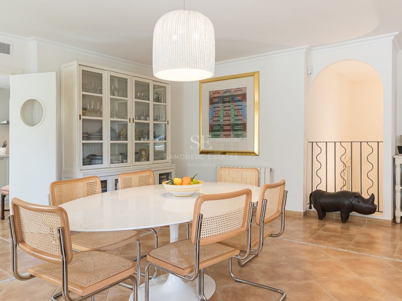 Bright dining room featuring an oval white table, rattan chairs, terracotta floors, and a large display cabinet.
