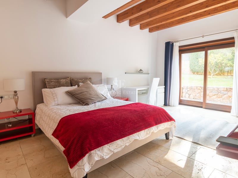 Ljust sovrum med röda detaljer, stengolv, takbjälkar i trä och skjutdörrar i glas mot trädgården.