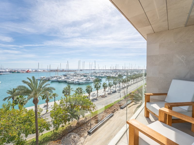 Modern balkong med trästolar och glasräcke med utsikt över en marina fylld med yachter och palmer.