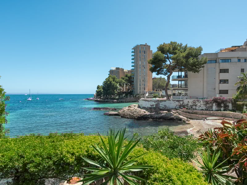 Turkos vik med klippig kust och moderna byggnader inramade av medelhavsgrönska under en klarblå himmel.