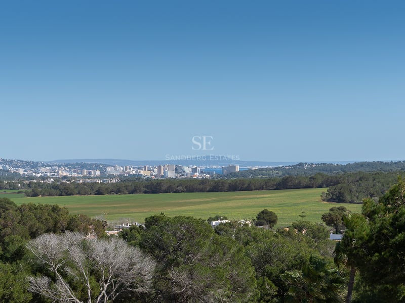 Upphöjd landskapsvy över gröna fält och tallskogar mot stadens silhuett och havet i fjärran.