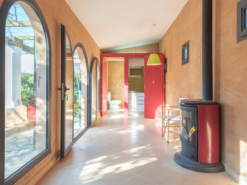 Solbelyst rum med terrakottaväggar, valvade glasdörrar, en röd braskamin och utsikt mot trädgården.