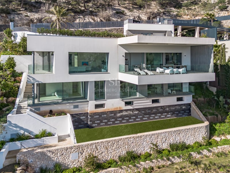 Villa blanche moderne de plusieurs étages avec de grandes baies vitrées et terrasses construite sur une colline rocheuse.