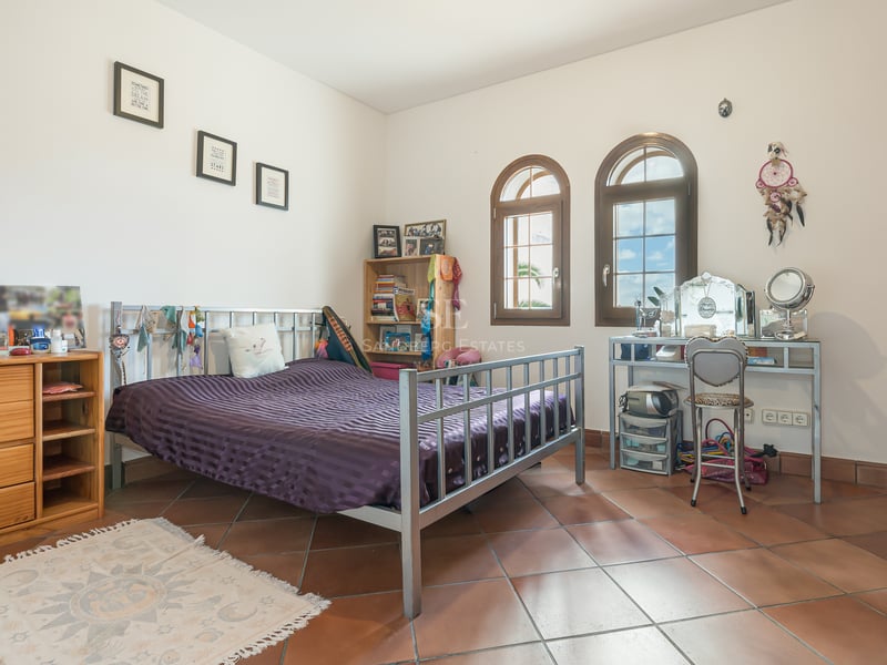 Bedroom featuring terracotta tiles, metal bed frame, arched wooden windows, and built-in wardrobes under natural light.
