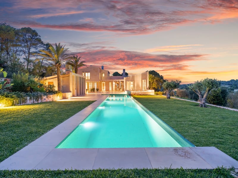 Longue piscine éclairée devant une villa moderne en pierre sous un ciel de coucher de soleil rose et orange.