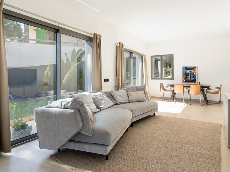 Ljust vardagsrum med grå soffa, stora glaspartier mot trädgården och matplats med läderstolar.