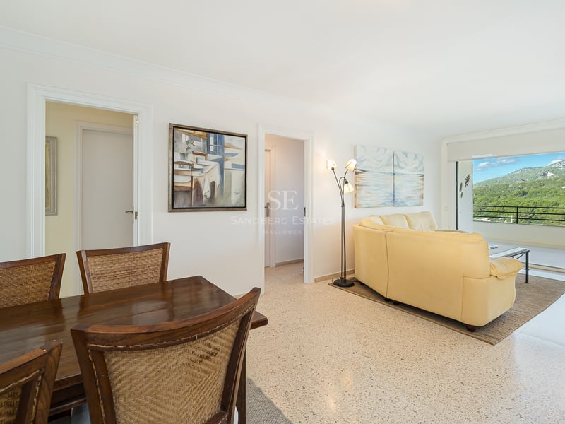 Open living space with a cream leather sofa, wooden dining set, and large glass doors revealing a mountain landscape.