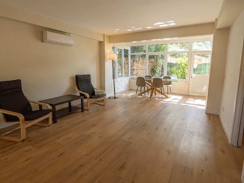 Salón con suelos de madera, grandes ventanales con vistas al jardín, mesa de comedor de cristal y dos sillones negros.