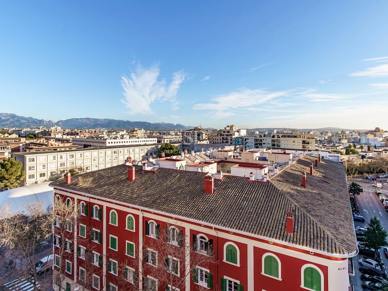 Vue en plongée d'une ville avec un bâtiment rouge, des rues et des montagnes au loin sous un ciel dégagé.